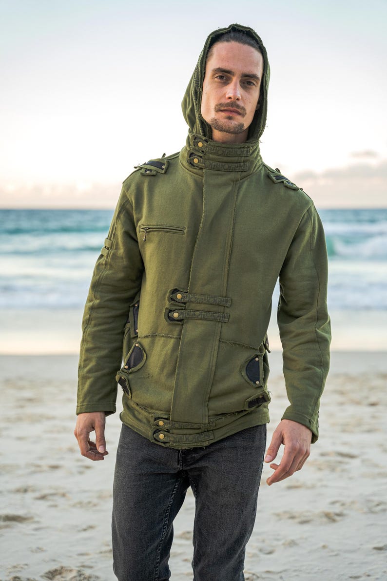 May include: An olive green hooded jacket with multiple button closures and decorative accents. The jacket features a high collar and a fitted silhouette. The person in the image is wearing dark gray jeans. The background is a beach with the ocean in the distance.