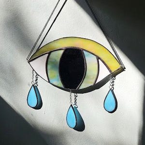 May include: Stained glass eye art piece with a yellow, white, and green iris, and a black pupil. The eye has a yellow border and three blue teardrop pendants hanging from the bottom. The piece is suspended by black strings.