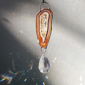May include: A natural agate slice suncatcher with a teardrop crystal pendant. The agate features bands of orange, brown, and white. The crystal reflects light, creating a rainbow effect. The suncatcher hangs from a black cord.