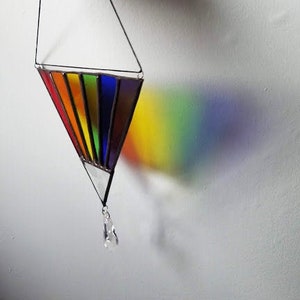 May include: A stained glass rainbow prism hanging from a black string with a crystal drop at the bottom. The prism casts a rainbow shadow on the white wall.
