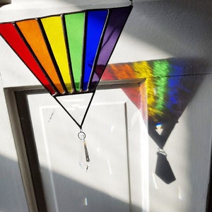 May include: A triangular stained glass window hanging from a white door. The window is made of colorful glass, with each panel a different color of the rainbow. A crystal prism hangs from the bottom of the window.