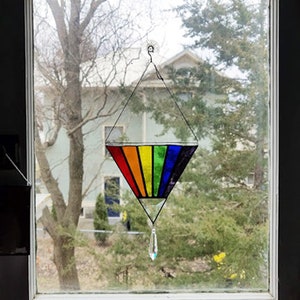 May include: A rainbow stained glass suncatcher hanging in a window. The suncatcher is shaped like a triangle with a crystal teardrop hanging from the bottom.