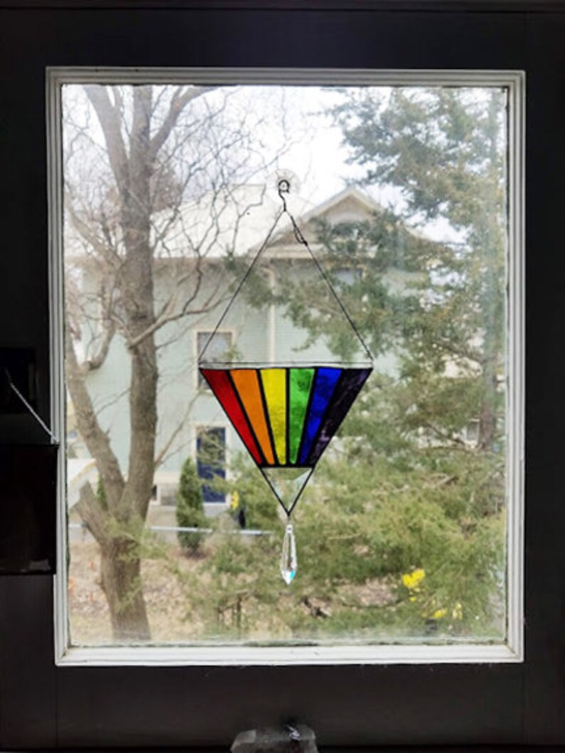 May include: A rainbow stained glass suncatcher hanging in a window. The suncatcher is shaped like a triangle with a crystal teardrop hanging from the bottom.