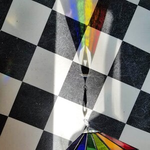 May include: A rainbow stained glass prism hanging from a black metal chain on a black and white checkered background.