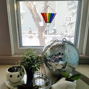 May include: A rainbow stained glass suncatcher hangs in a window. A silver disco ball sits on a table with a white ceramic planter with a smiley face design. A small potted plant sits in a white ceramic hand.