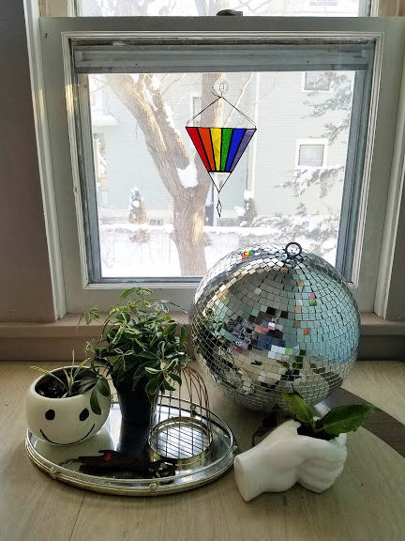 May include: A rainbow stained glass suncatcher hangs in a window. A silver disco ball sits on a table with a white ceramic planter with a smiley face design. A small potted plant sits in a white ceramic hand.