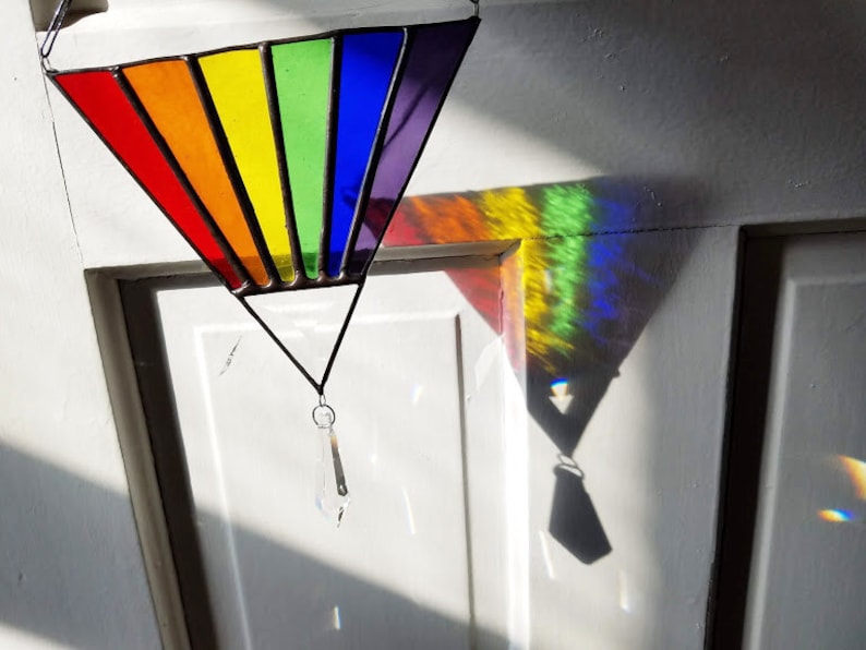 May include: A triangular stained glass window hanging from a white door. The window is made of colorful glass, with each panel a different color of the rainbow. A crystal prism hangs from the bottom of the window.