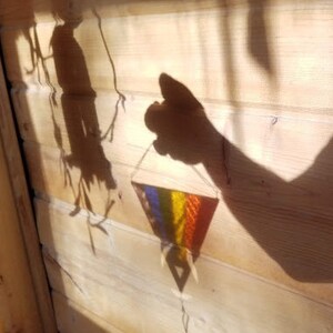 May include: A shadow of a hand holding a colorful woven triangle hanging from a string on a wooden wall.