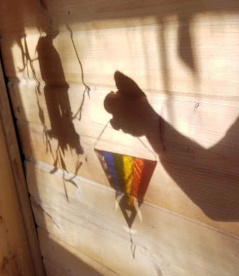 May include: A shadow of a hand holding a colorful woven triangle hanging from a string on a wooden wall.