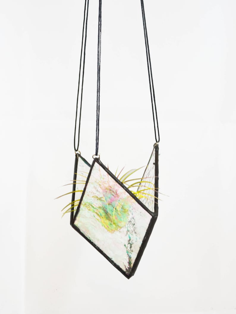 May include: A hanging geometric air plant terrarium. The glass is iridescent with a black metal frame. The terrarium is suspended by black cords and contains a small air plant with green leaves. The background is white.