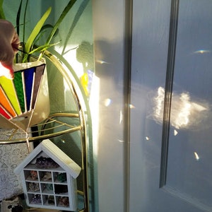 May include: A rainbow stained glass suncatcher hanging from a gold metal stand. The suncatcher is shaped like a triangle and has a variety of colors, including red, orange, yellow, green, blue, and purple. The suncatcher is casting rainbows on the wall behind it.