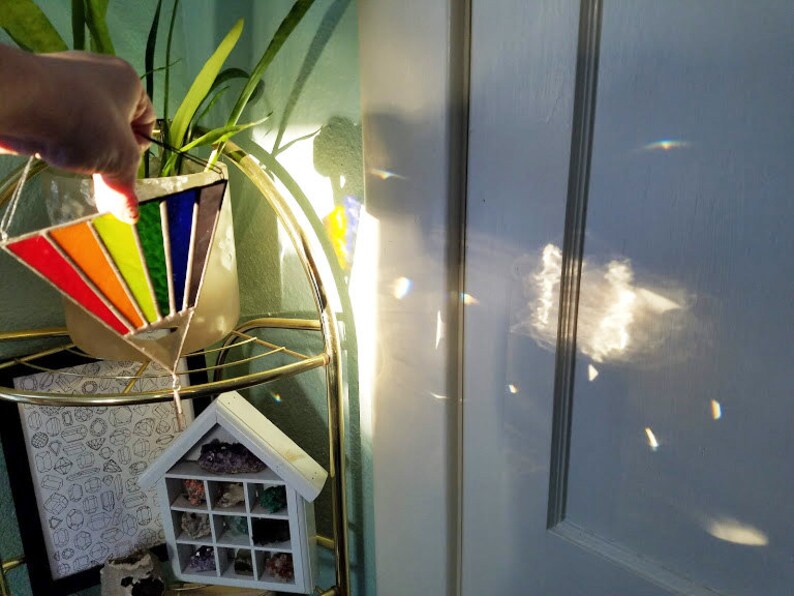 May include: A rainbow stained glass suncatcher hanging from a gold metal stand. The suncatcher is shaped like a triangle and has a variety of colors, including red, orange, yellow, green, blue, and purple. The suncatcher is casting rainbows on the wall behind it.