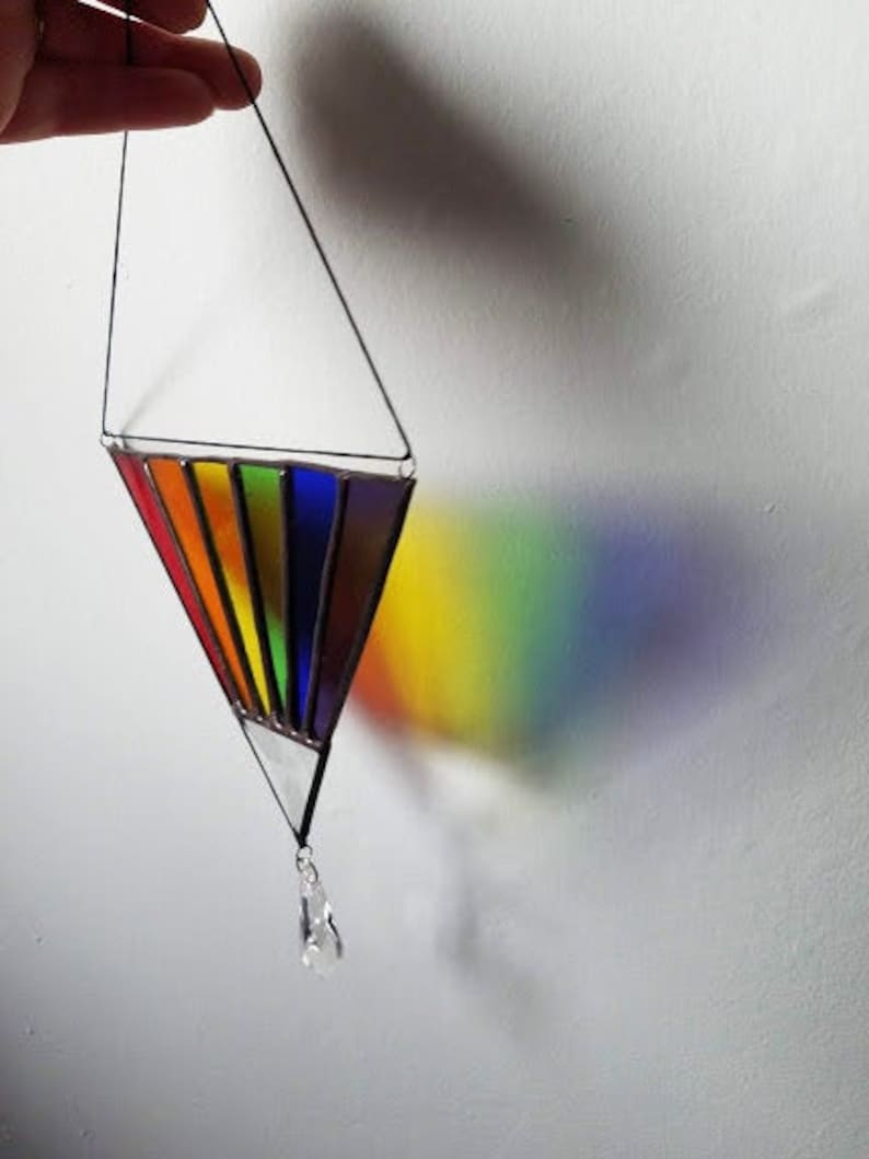 May include: A stained glass rainbow prism hanging from a black string with a crystal drop at the bottom. The prism casts a rainbow shadow on the white wall.