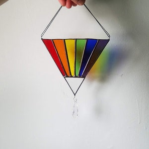 May include: A rainbow stained glass suncatcher with a crystal prism hanging from the bottom. The suncatcher is shaped like a diamond and is made of red, orange, yellow, green, blue, indigo, and violet glass.