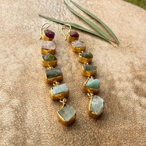 May include: A pair of long drop earrings featuring a vertical chain of raw, multi-coloured gemstones set in gold-toned metal. Each earring displays a sequence of stones in shades of deep red, pale pink, olive green, dark grey, and light blue.