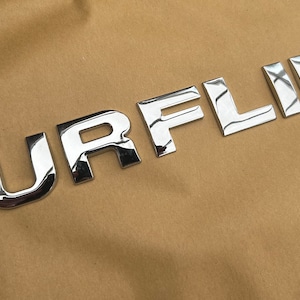 May include: Shiny chrome SURFLINE letters on a tan background. The letters are individual and appear to be 3D, reflecting light. The overall aesthetic suggests a product related to surfing or water sports.