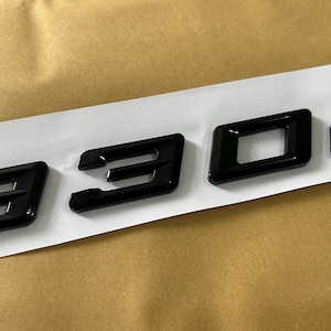 May include: Black 330d car badge with a glossy finish. The letters and numbers are arranged on a white strip, set against a tan background. The emblem is likely for a vehicle.
