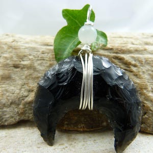 May include: A black obsidian crescent moon pendant with silver wire detailing and a small, round, white gemstone. The pendant is suspended from a silver wire loop and is set against a natural wood background.