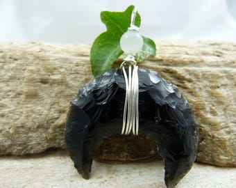 May include: A black obsidian crescent moon pendant with silver wire detailing and a small, round, white gemstone. The pendant is suspended from a silver wire loop and is set against a natural wood background.