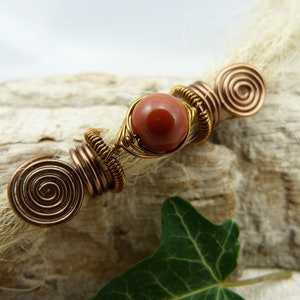 May include: A brown and gold wire wrapped bead with a red stone in the center. The bead is attached to a strand of white hair.