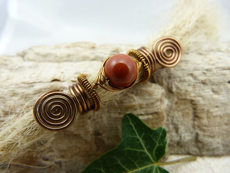 May include: A brown and gold wire wrapped bead with a red stone in the center. The bead is attached to a strand of white hair.