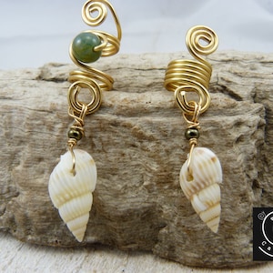 Può includere: Un paio di orecchini dorati con una pietra verde e una conchiglia bianca che pendono da ciascuno. Gli orecchini sono fatti di filo metallico e sono progettati per avvolgersi attorno all'orecchio.