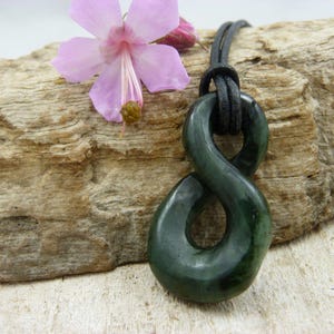 Könnte beinhalten: Ein dunkelgrüner Jade-Anhänger in Unendlichkeitsform, auf einer schwarzen Kordel. Der Anhänger ist glatt und poliert, mit leichtem Glanz. Eine rosa Blume liegt auf einem Stück Treibholz im Hintergrund.
