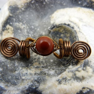 May include: A copper wire hair wrap with a red gemstone bead in the center. The wire is twisted into a spiral shape on each end.