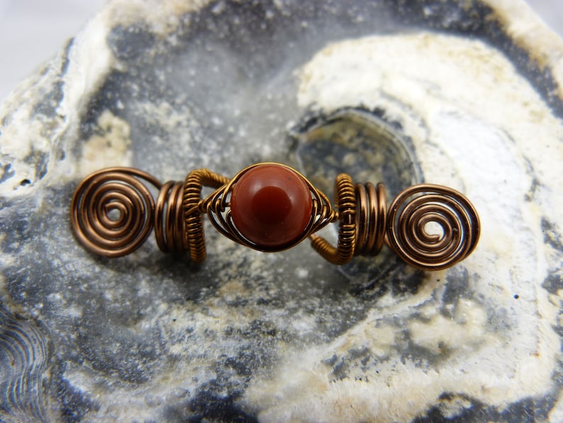 May include: A copper wire hair wrap with a red gemstone bead in the center. The wire is twisted into a spiral shape on each end.