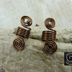 Könnte beinhalten: Zwei Haarschmuckspiralen aus Kupferdraht, jede mit einer kleinen Perle oben. Die Spiralen liegen auf einer Holzoberfläche.