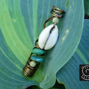 Peut inclure: Accessoire pour cheveux artisanal avec un coquillage blanc, des perles turquoise et un design en spirale. L'accessoire est fabriqué avec du fil de couleur bronze et est présenté sur une grande feuille verte. Un petit logo est visible en bas à droite.