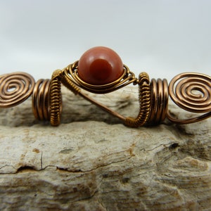 May include: A copper wire hair wrap with a spiral design on each end and a red-brown gemstone in the center.