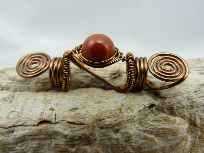 May include: A copper wire hair wrap with a spiral design on each end and a red-brown gemstone in the center.