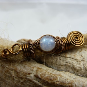 Puede incluir: Una cuenta de rasta de alambre de cobre con una cuenta de piedra lunar blanca en el centro. La cuenta está envuelta en alambre de cobre y tiene un diseño en espiral en un extremo.