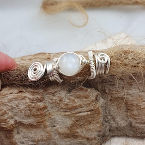 Könnte beinhalten: Ein silberfarbenes Haaraccessoire aus Draht mit einem zentralen, runden, weißen Edelstein. Der Draht ist kunstvoll um den Edelstein gewickelt und weist spiralförmige Designs auf beiden Seiten auf. Das Accessoire wird auf einer strukturierten, braunen Oberfläche gezeigt.
