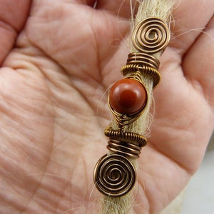 May include: A brown gemstone bead with copper wire wrapped around it, with two copper wire spirals on either end. The bead and wire are attached to a strand of light brown hair.