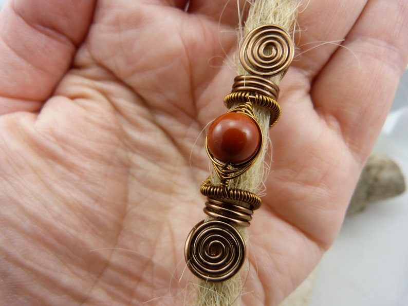May include: A brown gemstone bead with copper wire wrapped around it, with two copper wire spirals on either end. The bead and wire are attached to a strand of light brown hair.