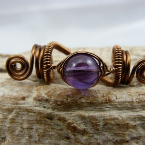 May include: Copper-coloured hair accessory with a central purple gemstone. The wire-wrapped design features spiral accents on each end and a circular frame around the gemstone. The accessory is designed for hair styling.
