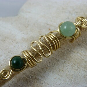 May include: Gold wire wrapped around a brown hair strand with two green gemstone beads.