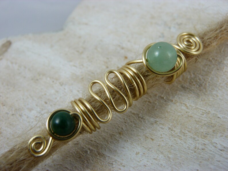 May include: Gold wire wrapped around a brown hair strand with two green gemstone beads.