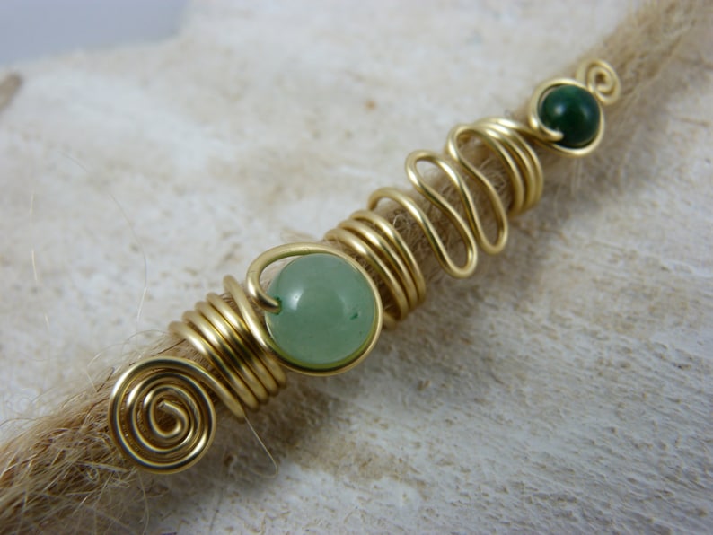 May include: Gold wire wrapped dreadlock beads with green gemstone accents. The beads are wrapped in a spiral pattern and feature two round green stones.