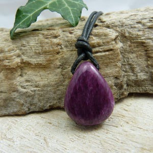 Könnte beinhalten: Ein Anhänger aus violettem Edelstein an einem schwarzen Lederband. Der Anhänger ist tropfenförmig und hat eine glatte, polierte Oberfläche.