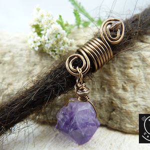 Könnte beinhalten: Eine Dreadlock-Perle aus Kupferdraht mit einem violetten Amethyst-Kristall-Anhänger. Die Perle ist um eine braune Dreadlock gewickelt.