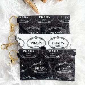 May include: Three small black and white Prada wallets with gold keyrings. The wallets feature a repeating logo pattern and a snap closure. One wallet is white with a black logo, and the other two are black with a white logo.