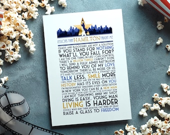 May include: A white rectangular print featuring a blue and gold graphic of a star and city skyline above the text LESSONS THAT HAMILTON TAUGHT ME. The print contains various black and blue lyrics from the musical Hamilton in a vertical layout.