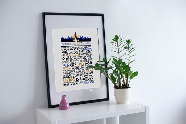 May include: A framed print with a white background and black frame. The print features a blue and gold design with the text "LESSONS THAT HAMILTON TAUGHT ME" and a list of quotes from the musical Hamilton.