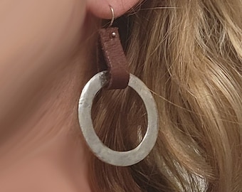 May include: A pair of silver hoop earrings with a brown leather strap hanging from each hoop.