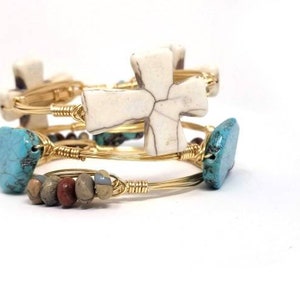 May include: Set of four gold wire bracelets with turquoise and white cross charms. The bracelets are stacked on top of each other.