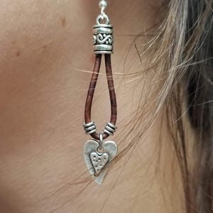 May include: A silver-toned earring with a heart-shaped pendant. The earring features a brown leather cord and decorative silver accents. The heart pendant has a smaller heart cutout design. The earring is shown against a blurred background.