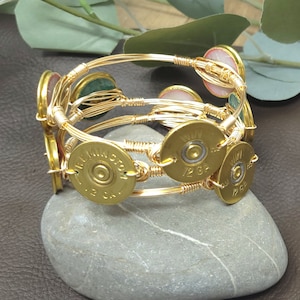 May include: Gold wire-wrapped bracelet featuring brass bullet casings and colorful gemstone accents. The bracelet is made with multiple strands of gold wire, creating a layered effect. The bullet casings are stamped with "Remington 12 GA".